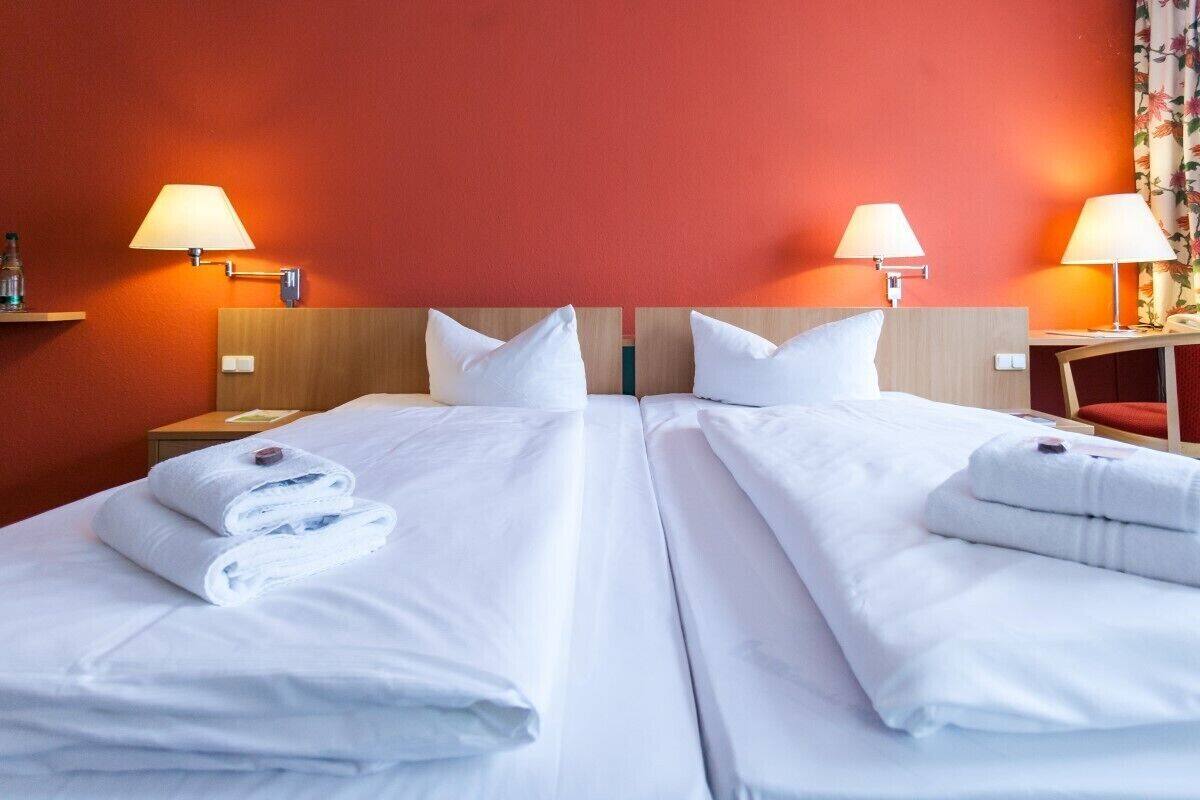 Quarto de hotel com duas camas, cada uma com roupa de cama e almofadas brancas, duas luminárias de cabeceira e uma pequena mesa com abajur.