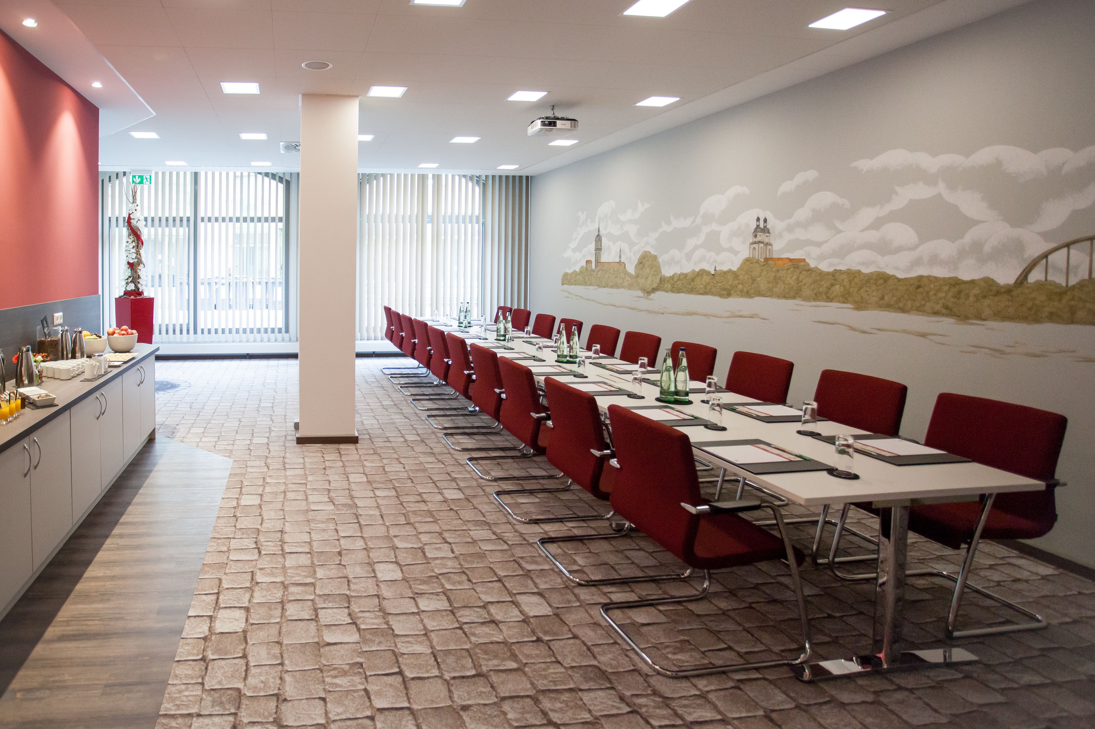 Ein Konferenzraum mit roten Stühlen und Tischen, einem Fliesenboden und einem Wandgemälde einer Stadtansicht.