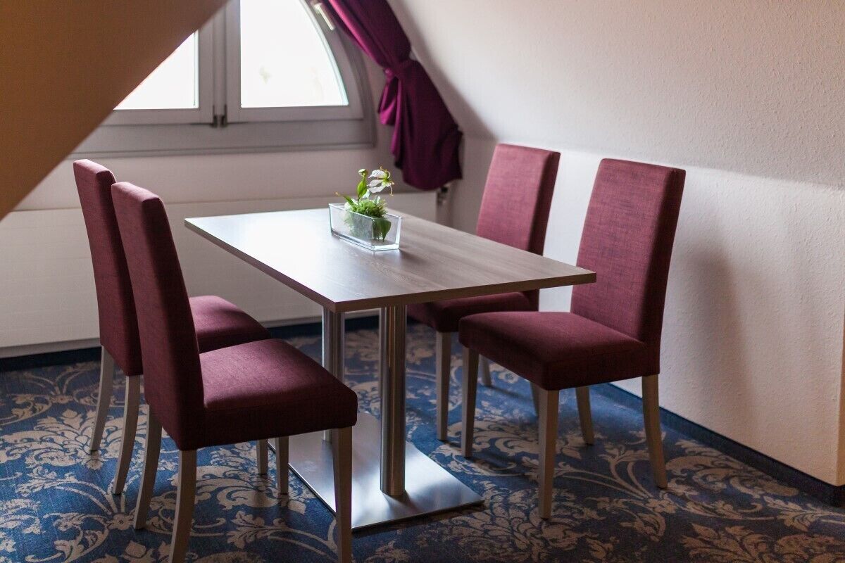 A dining area with a table and four red upholstered chairs. There is a small plant on the table, and the room has patterned carpet and a window with curtains.