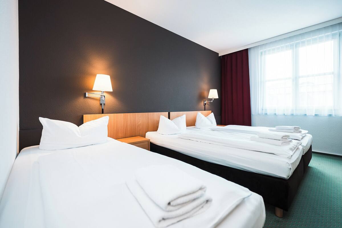 Quarto de hotel com duas camas, cada uma com lençóis e almofadas brancos, uma cabeceira de madeira e uma mesa de cabeceira com abajur.