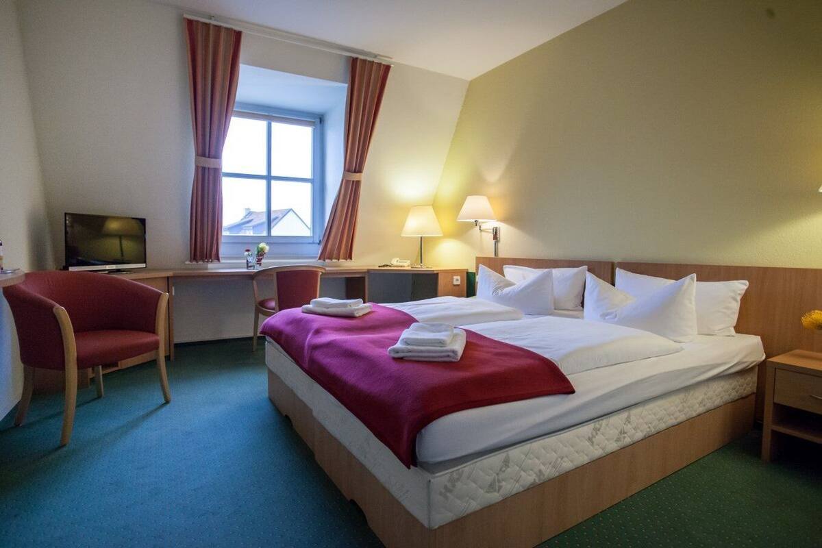 Quarto de hotel com cama, escrivaninha, cadeira, televisão e janela com cortinas.