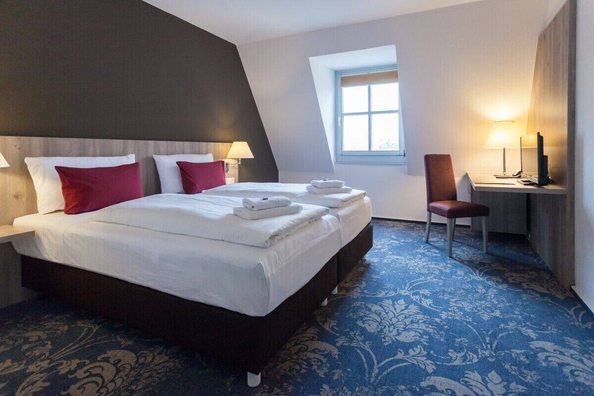 Quarto de hotel com uma cama grande, duas mesas de cabeceira com luminárias, uma escrivaninha com cadeira e uma janela.