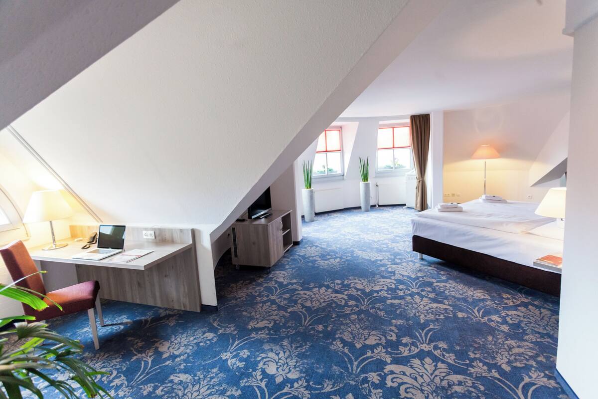 Quarto de hotel com escrivaninha, computador e cadeira, cama com luminária e carpete estampado azul.