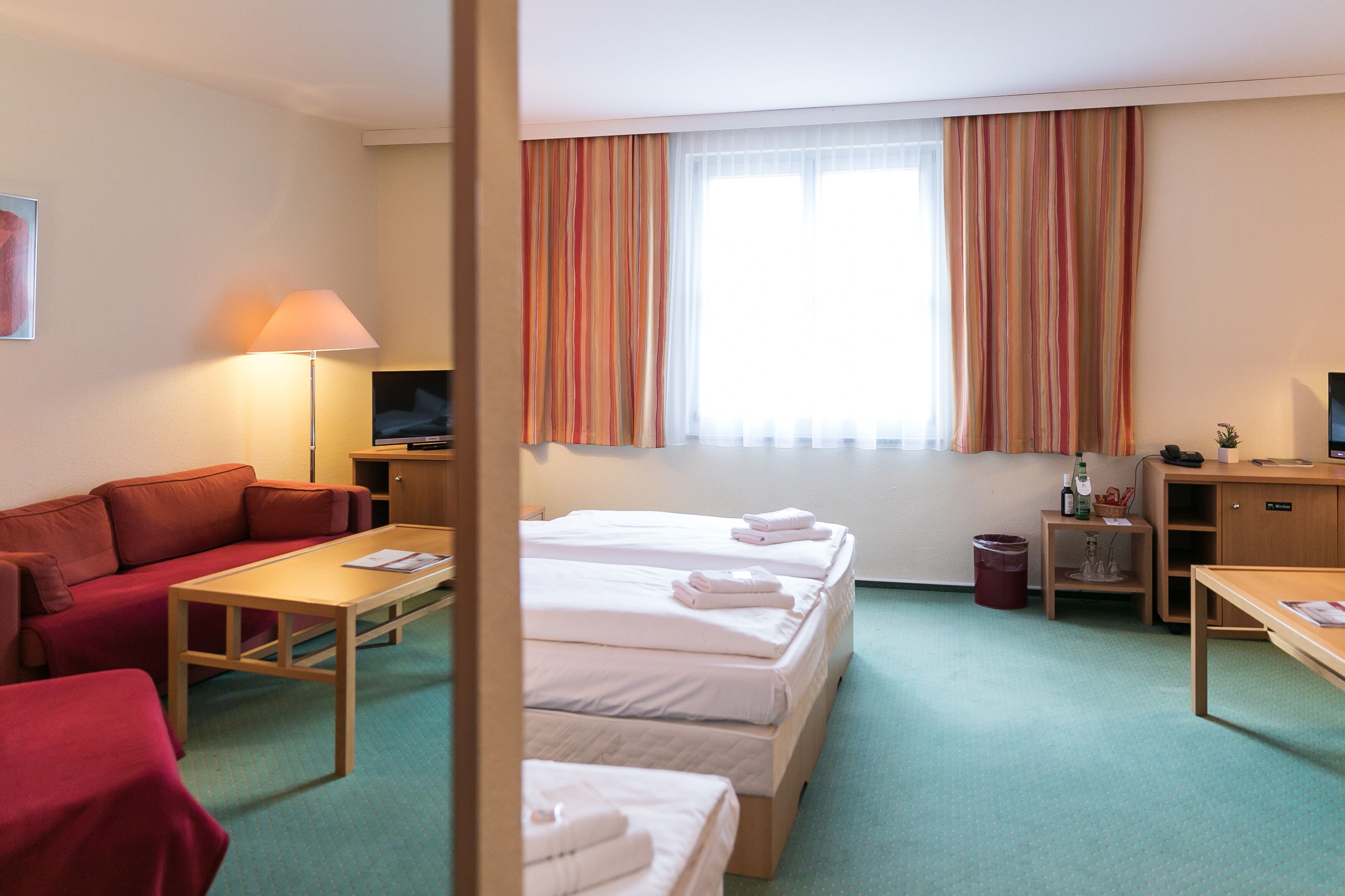 A hotel room with a sofa, two beds, a desk, a TV, and a window with curtains.