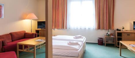 A hotel room with a sofa, two beds, a desk, a TV, and a window with curtains.