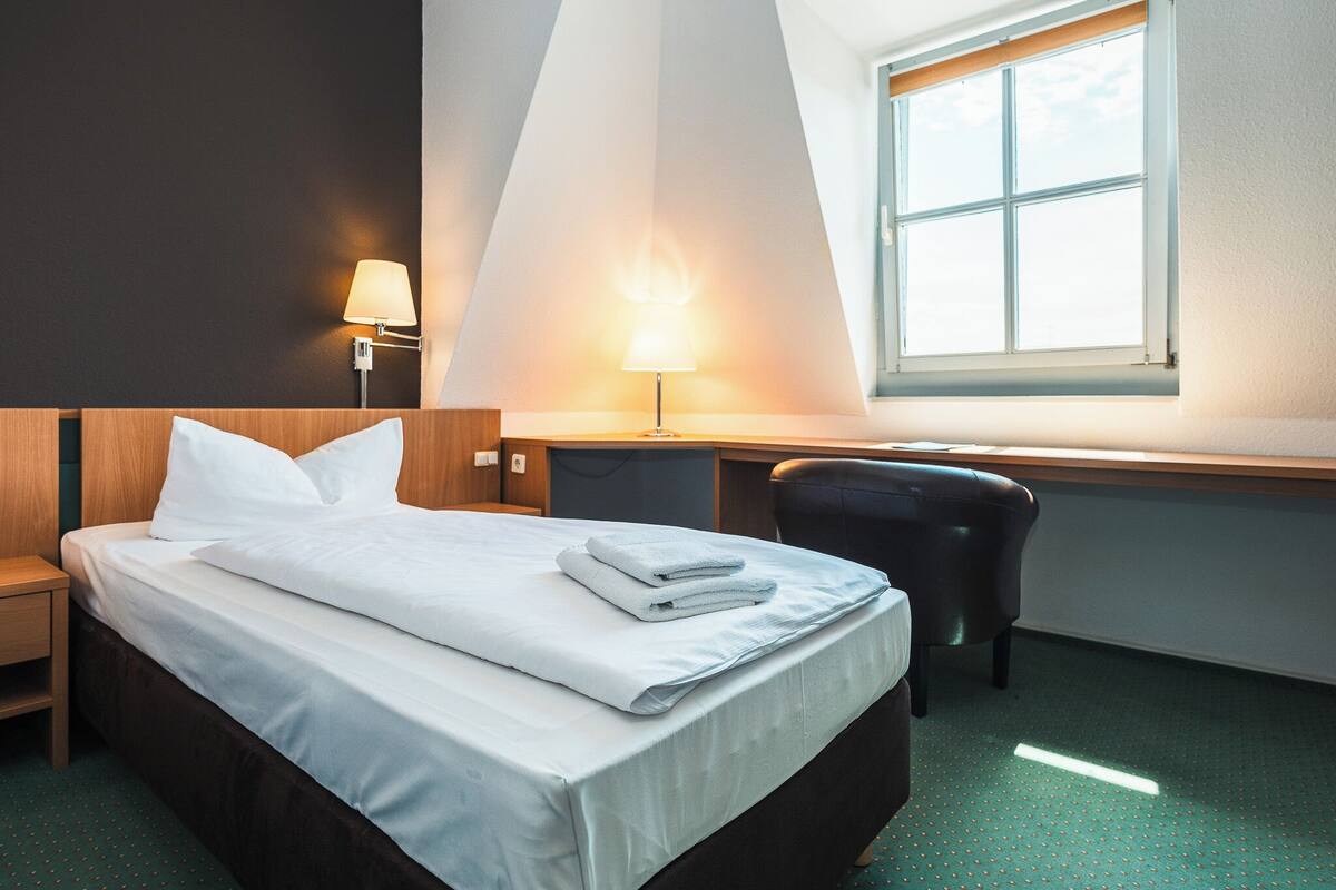 Quarto de hotel com uma cama bem arrumada, uma escrivaninha, uma cadeira, um abajur e uma janela.