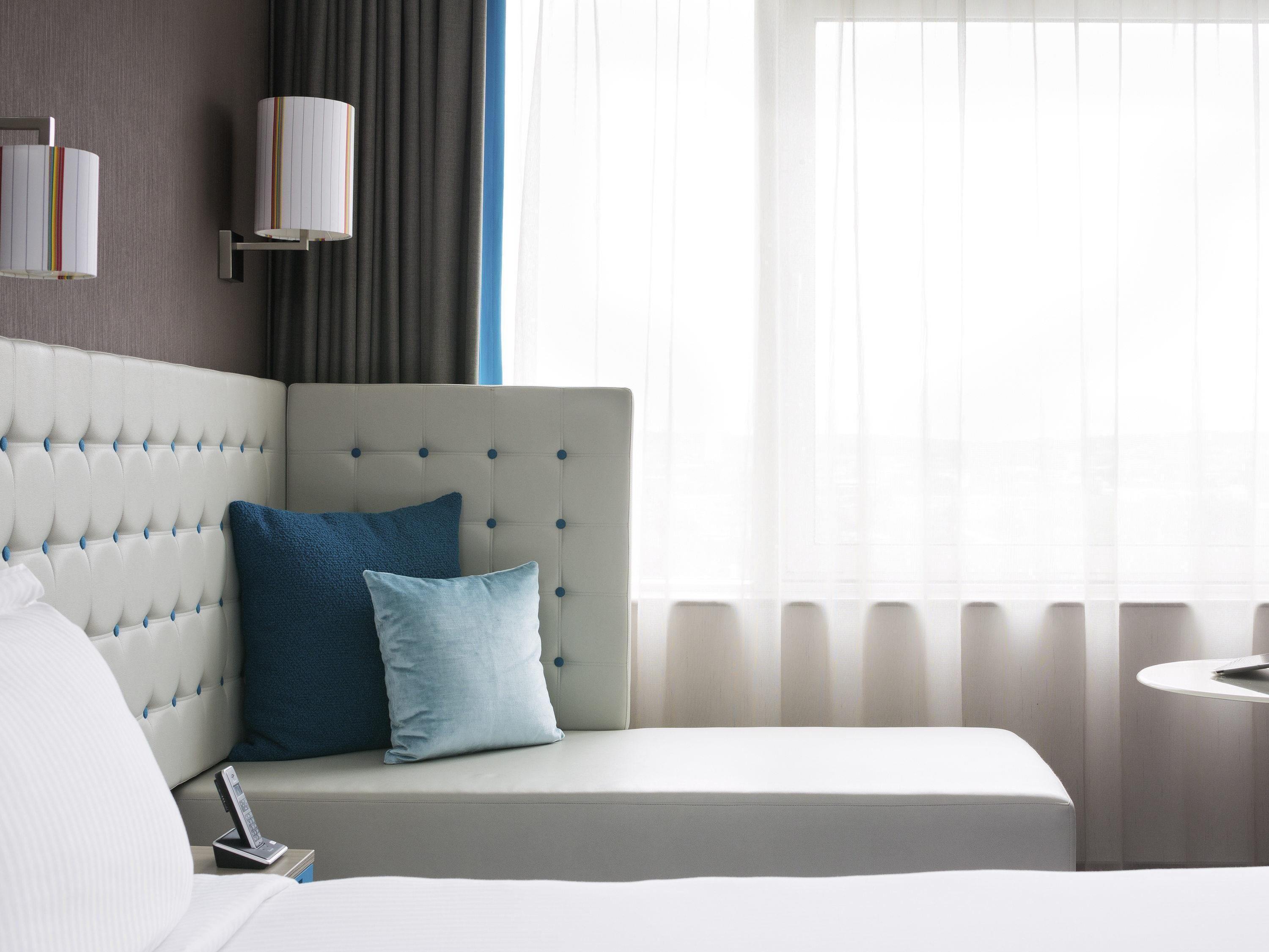 Una habitación de hotel con un cabecero capitonado, una mesita de noche con lámpara y una ventana con cortinas traslúcidas.
