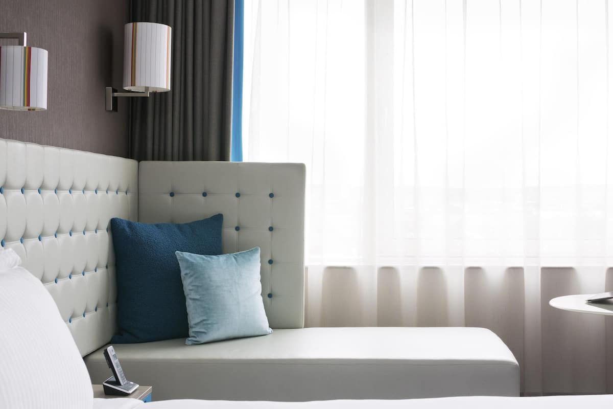 A hotel room with a tufted headboard, a bedside table with a lamp, and a window with sheer curtains.