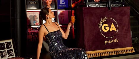 A woman in a sequined dress sitting on a stage with a microphone stand, holding a drink.
