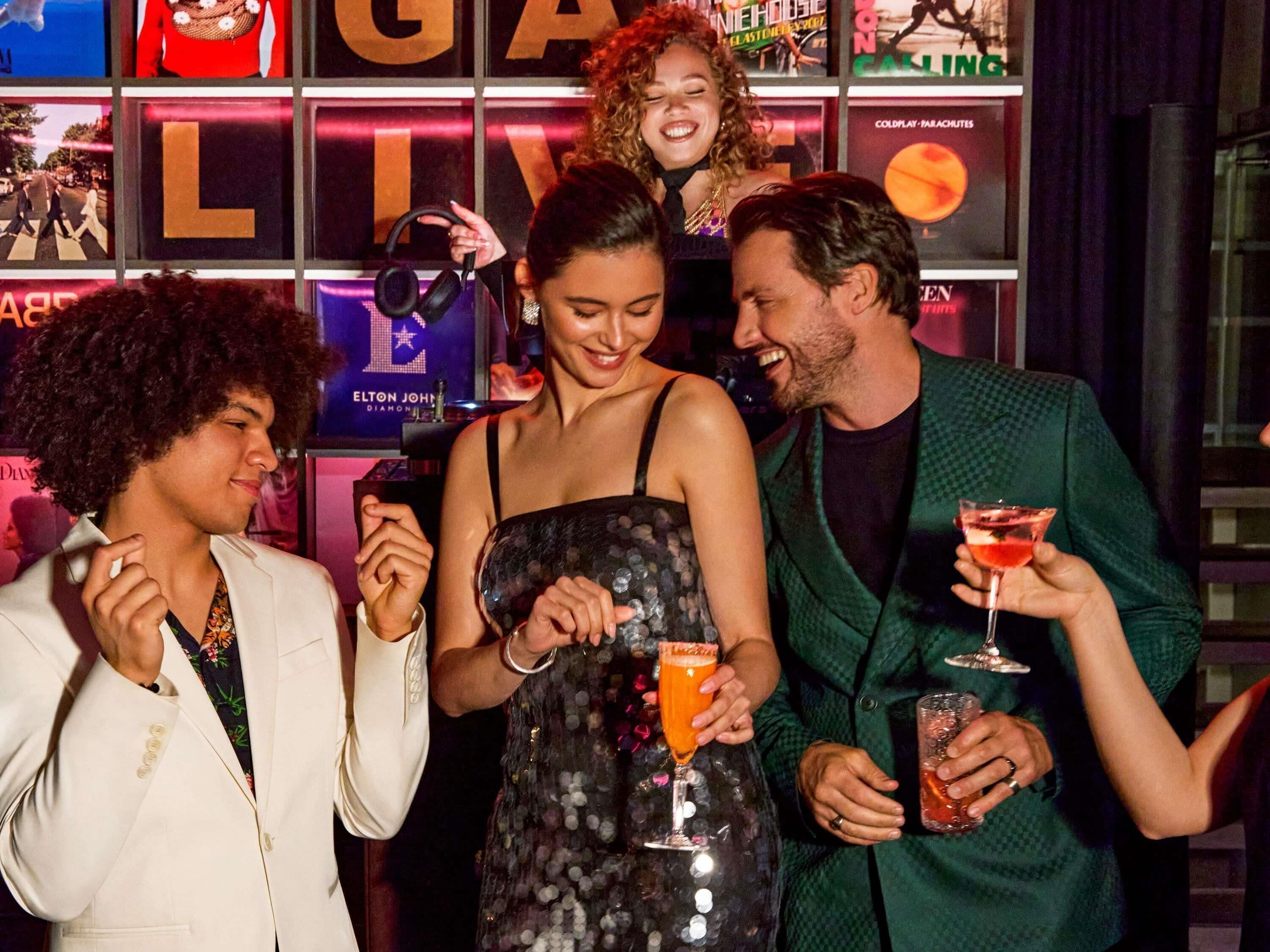 A group of people at a social event with a backdrop featuring a large illuminated 'G' and 'LIVE' sign, suggesting a lively atmosphere.