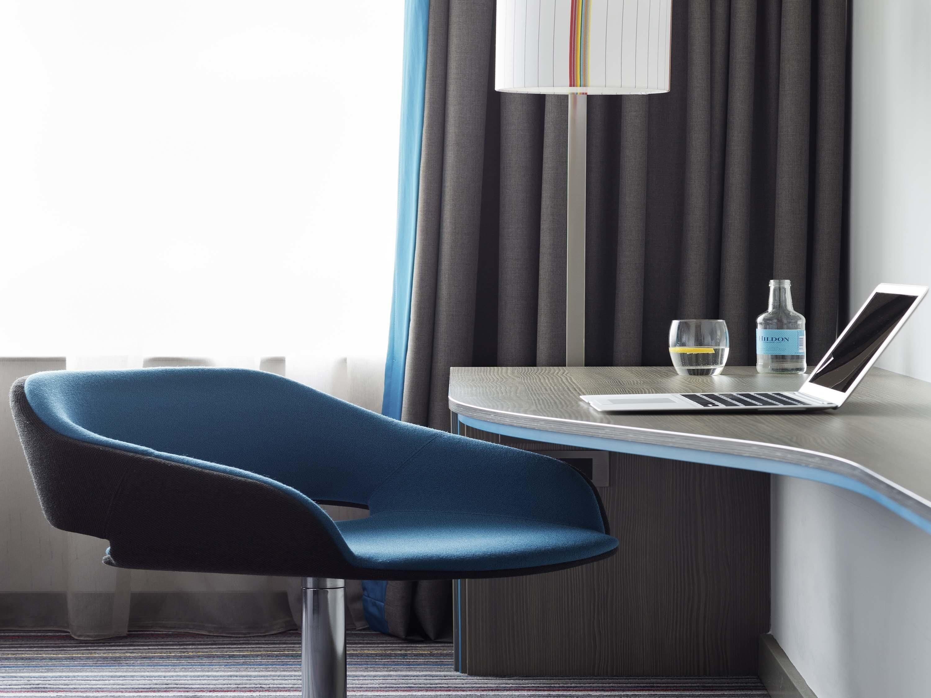 Une chambre d’hôtel moderne avec un bureau incurvé, un ordinateur portable, une bouteille d’eau et un verre.