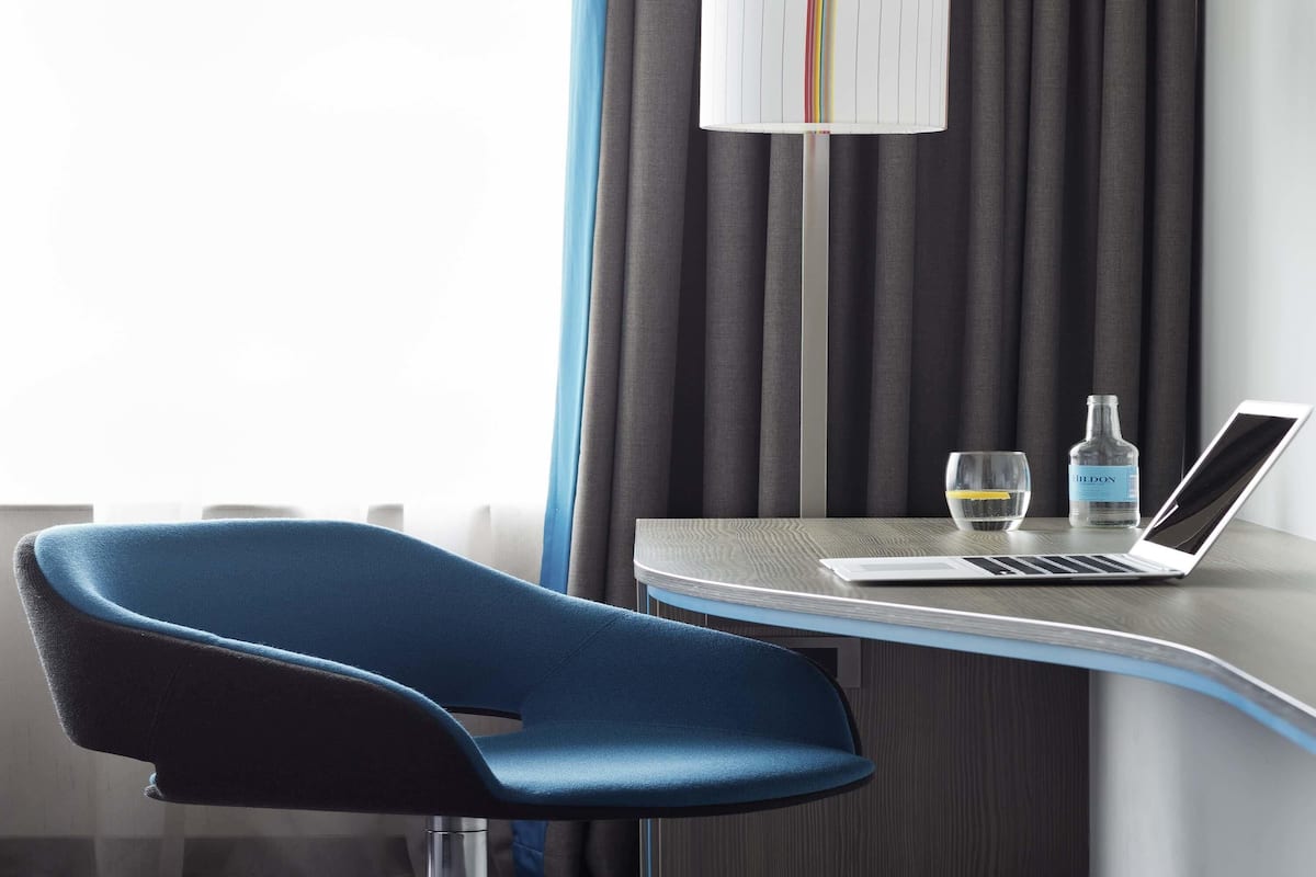 Une chambre dâhĂŽtel moderne avec un bureau incurvĂ©, un ordinateur portable, une bouteille dâeau et un verre.