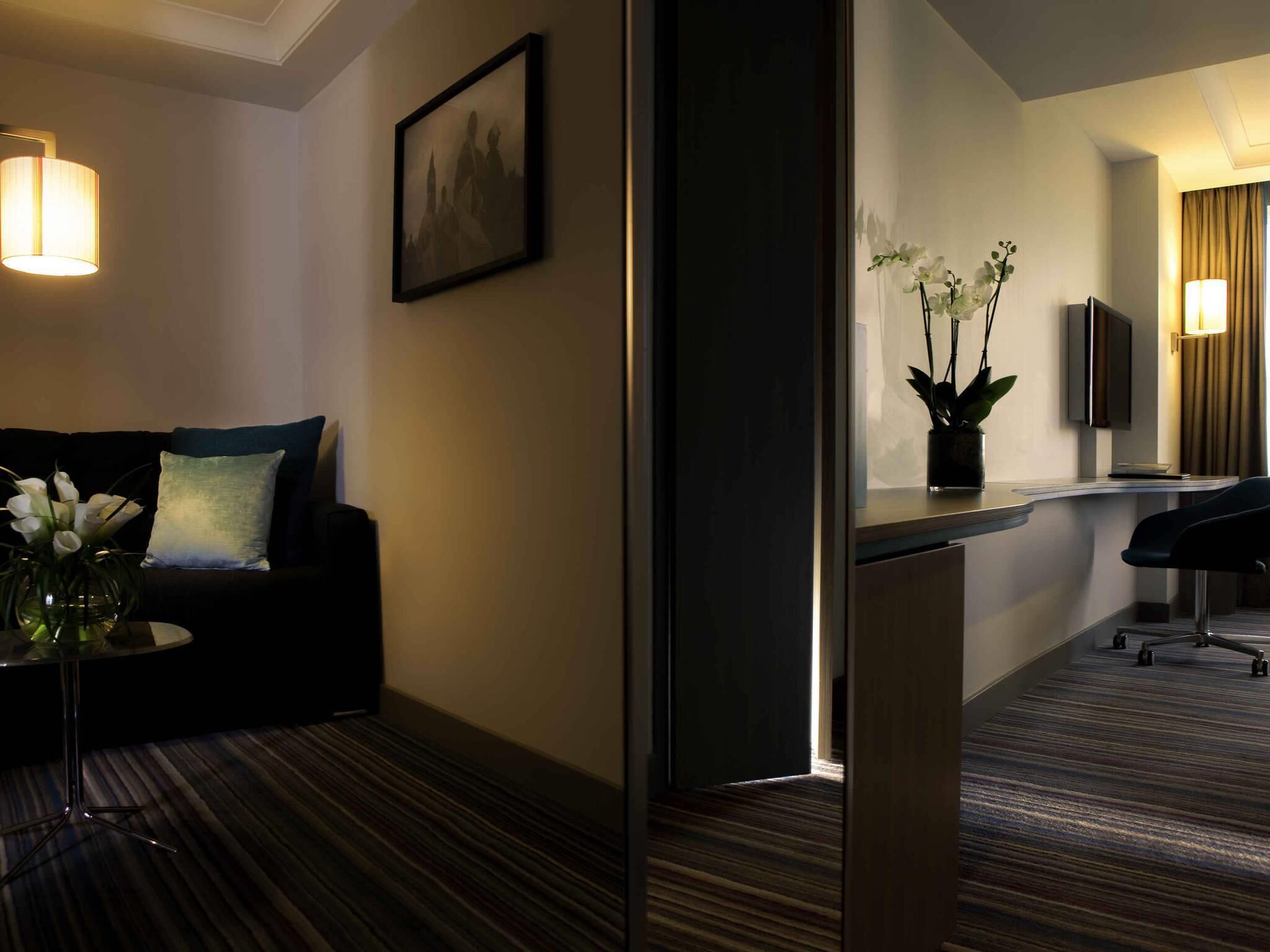 Habitación de hotel con sofá, una mesita con un florero, un escritorio con silla, un espejo y un televisor.