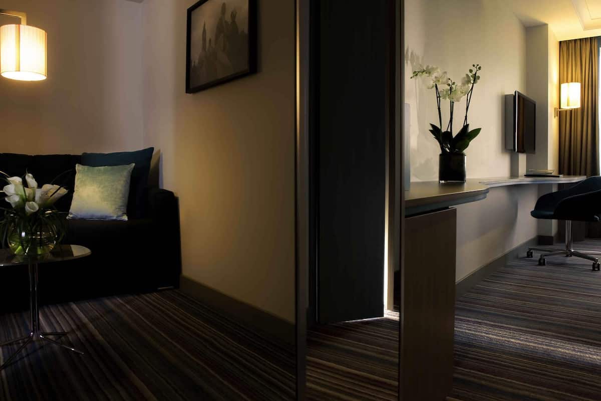 Habitación de hotel con sofá, una mesita con un florero, un escritorio con silla, un espejo y un televisor.