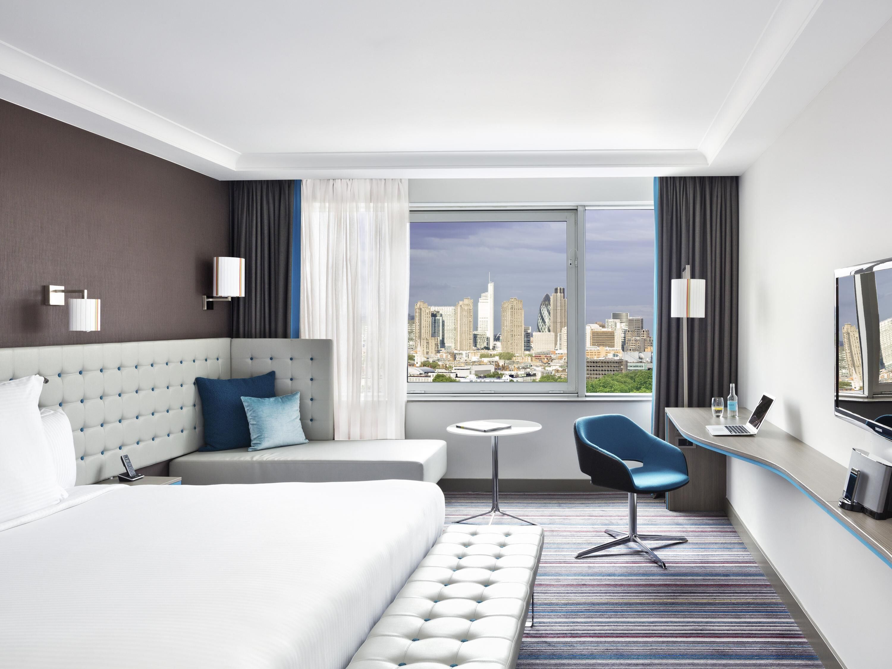 Habitación de hotel con cama, escritorio, silla, mesita pequeña y un amplio ventanal con vistas a la ciudad.