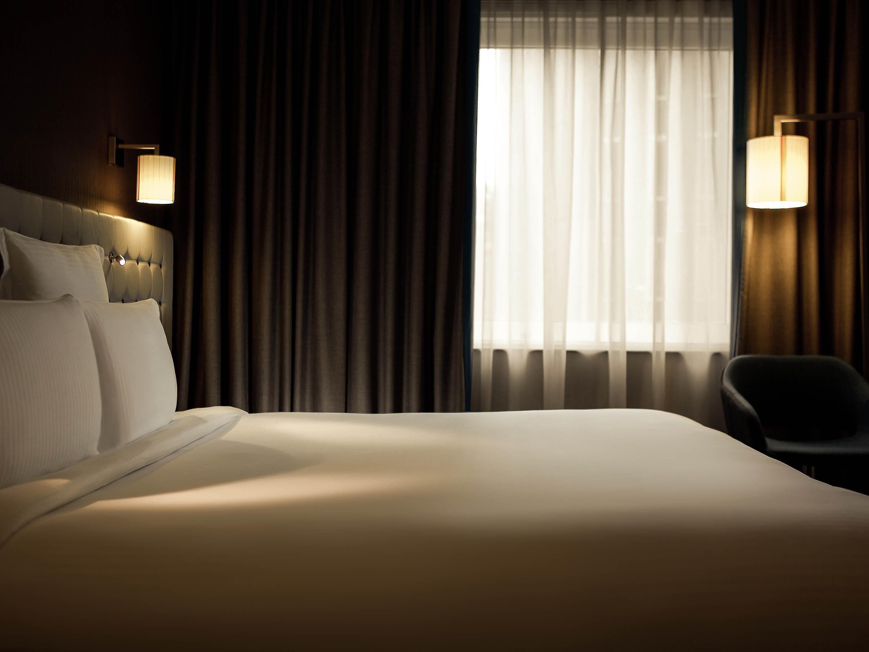 A hotel room with a large bed, two wall-mounted lamps, a chair, and a window with curtains.