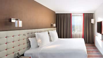 Ein Hotelzimmer mit einem großen Bett, einem roten Nachttisch, einem an der Wand befestigten Fernseher und einem Fenster mit Vorhängen.