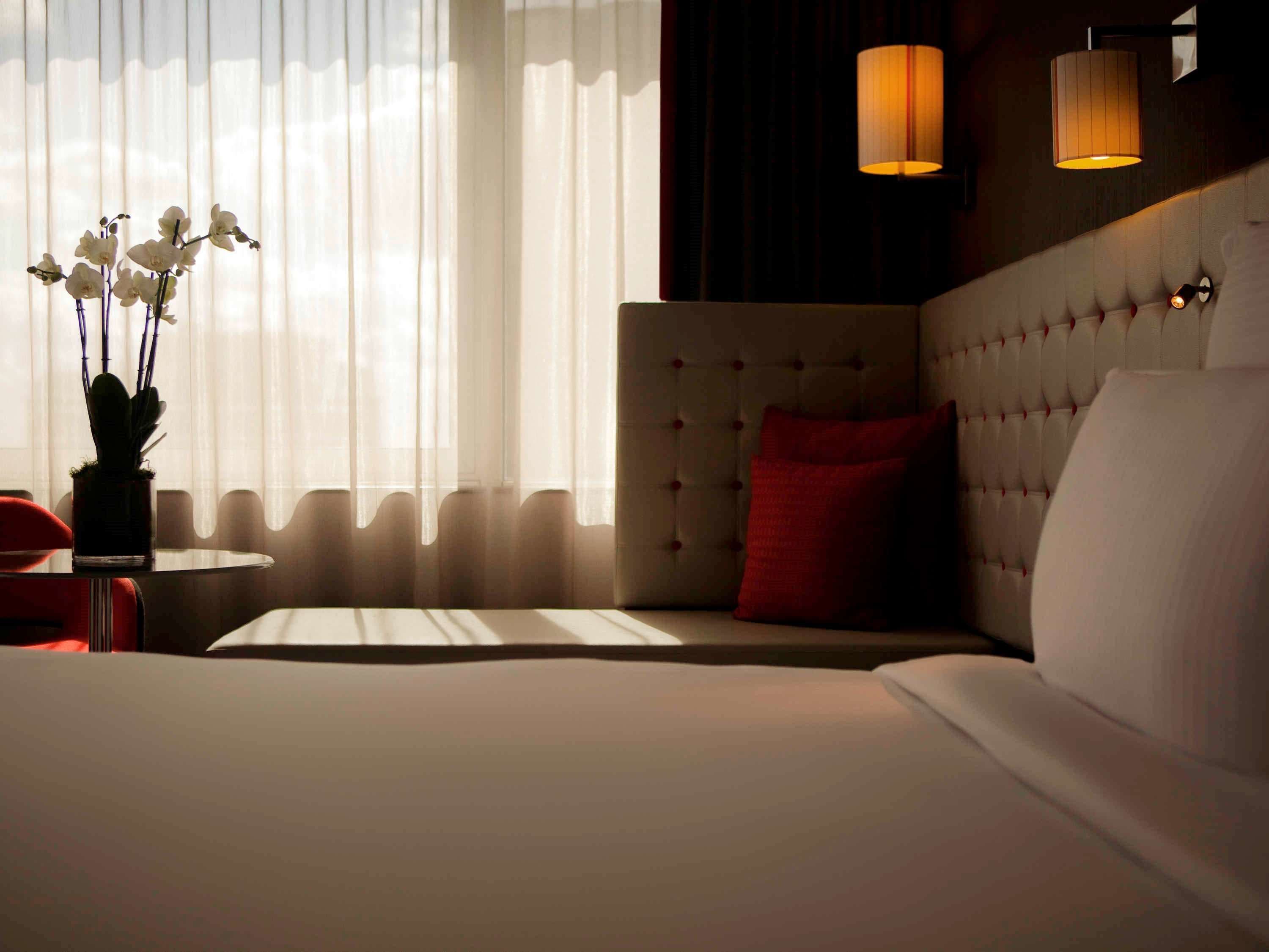 Ein Hotelzimmer mit einem großen Bett, einem roten Sessel, einem kleinen Tisch und einer Vase mit weißen Blumen.