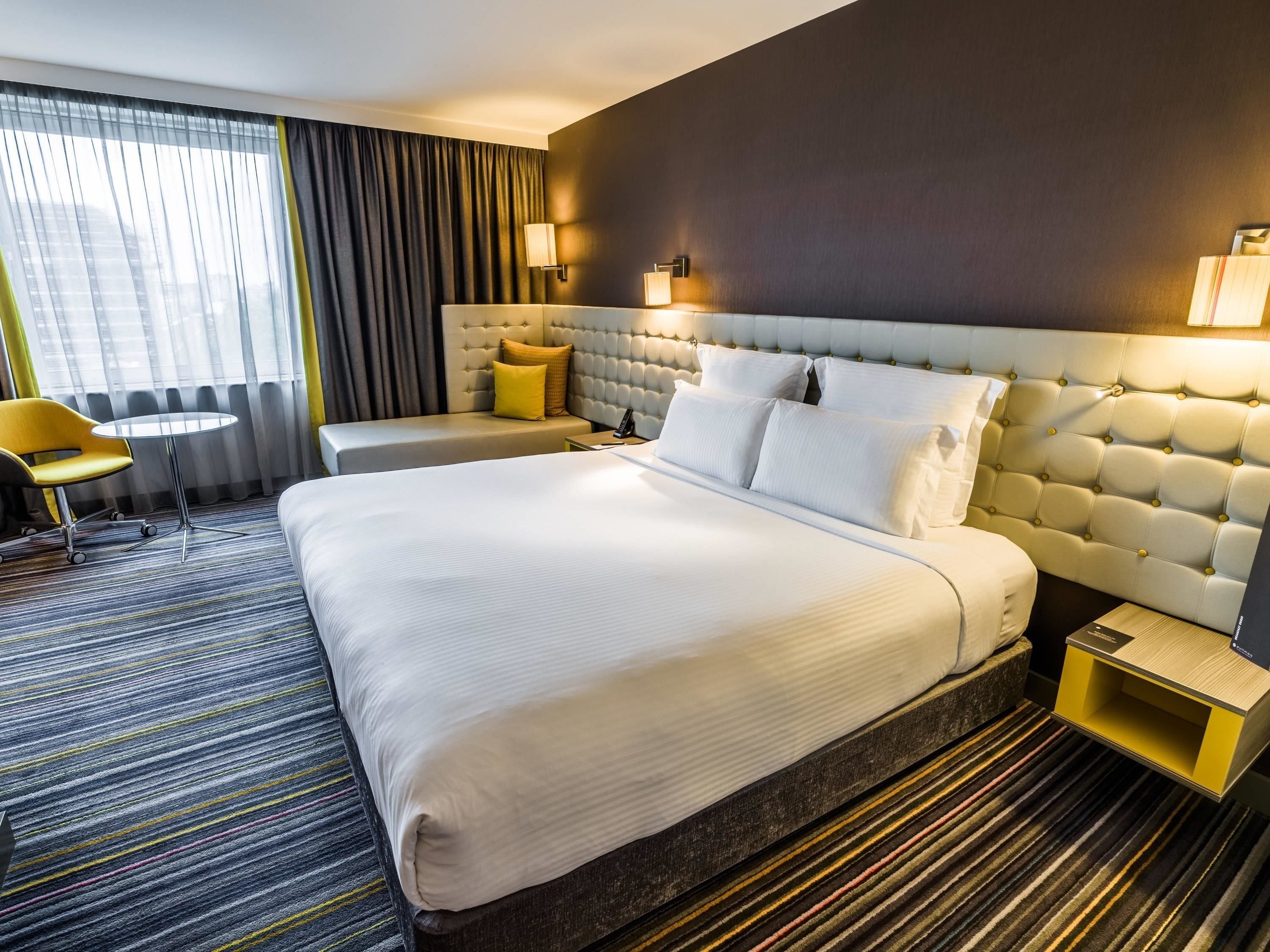 A hotel room with a large bed, a bedside table, a chair, a small round table, and a view of the city through the window.