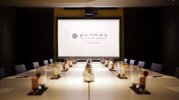 A conference room with a long table set for a meeting, a screen displaying the Pullman Hotels and Resorts logo, and a dark-colored wall.