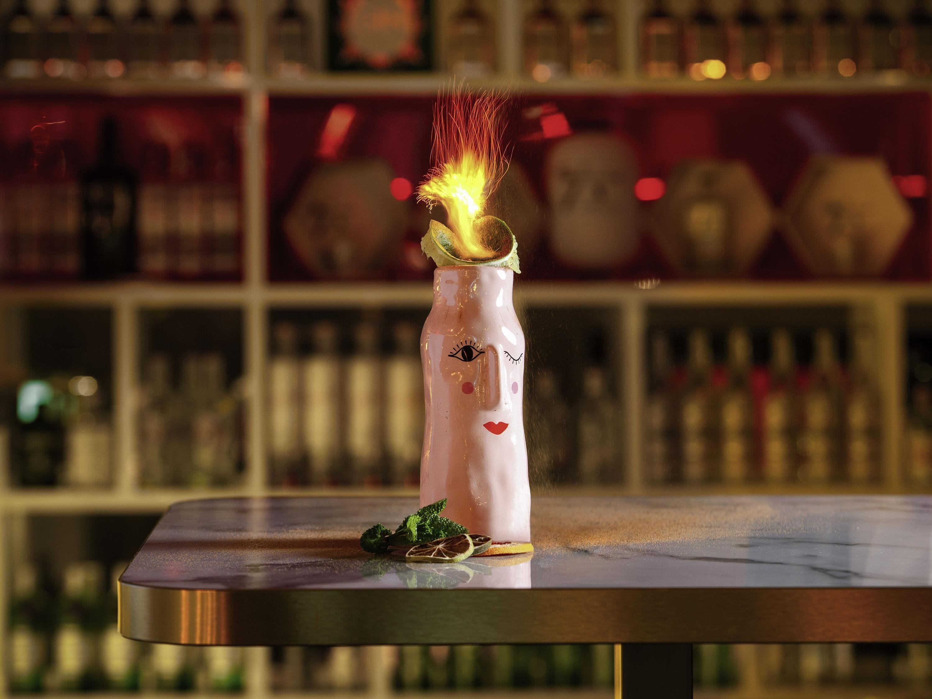 Eine Flasche mit Gesichtsmotiv und Sektglaspunkt auf einem Tisch, verschwommener Hintergrund mit Regalen.