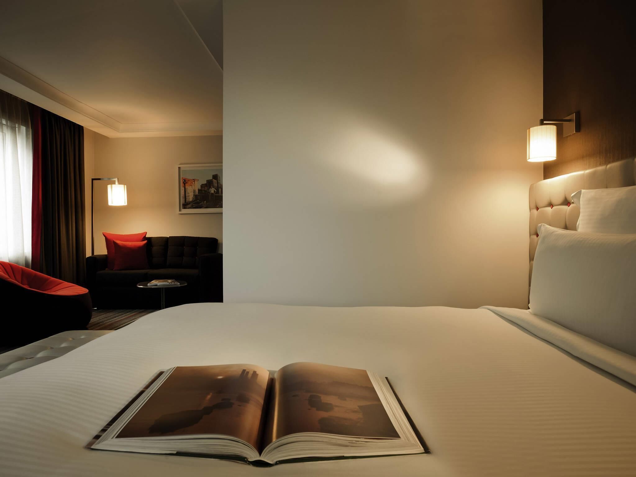 Una habitación de hotel con una cama grande, un sillón de lectura, una mesita pequeña y una mesita de noche.