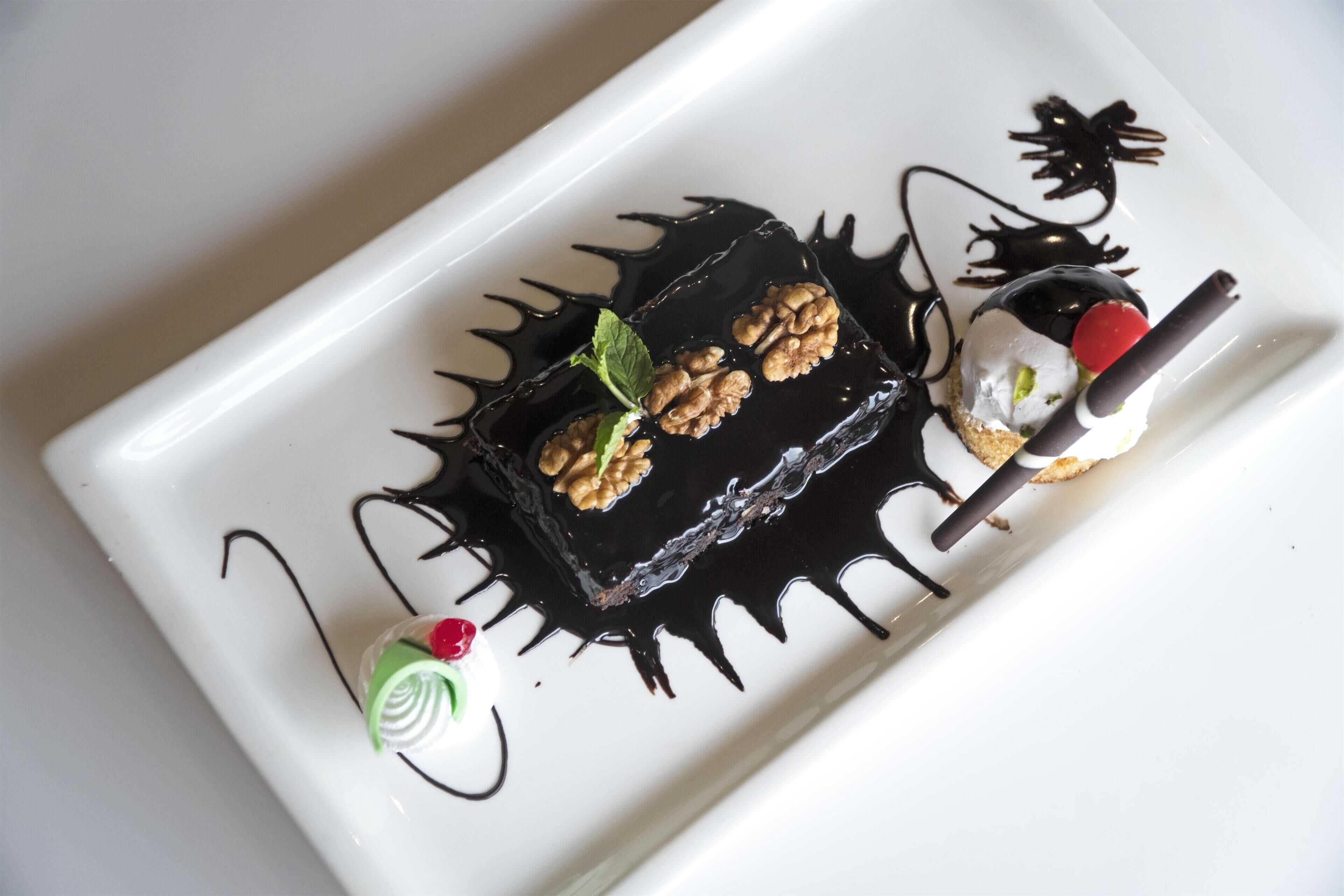 Un plato de postre con un cuadrado cubierto de chocolate, una bola de helado con cereza y un pequeño remolino verde.