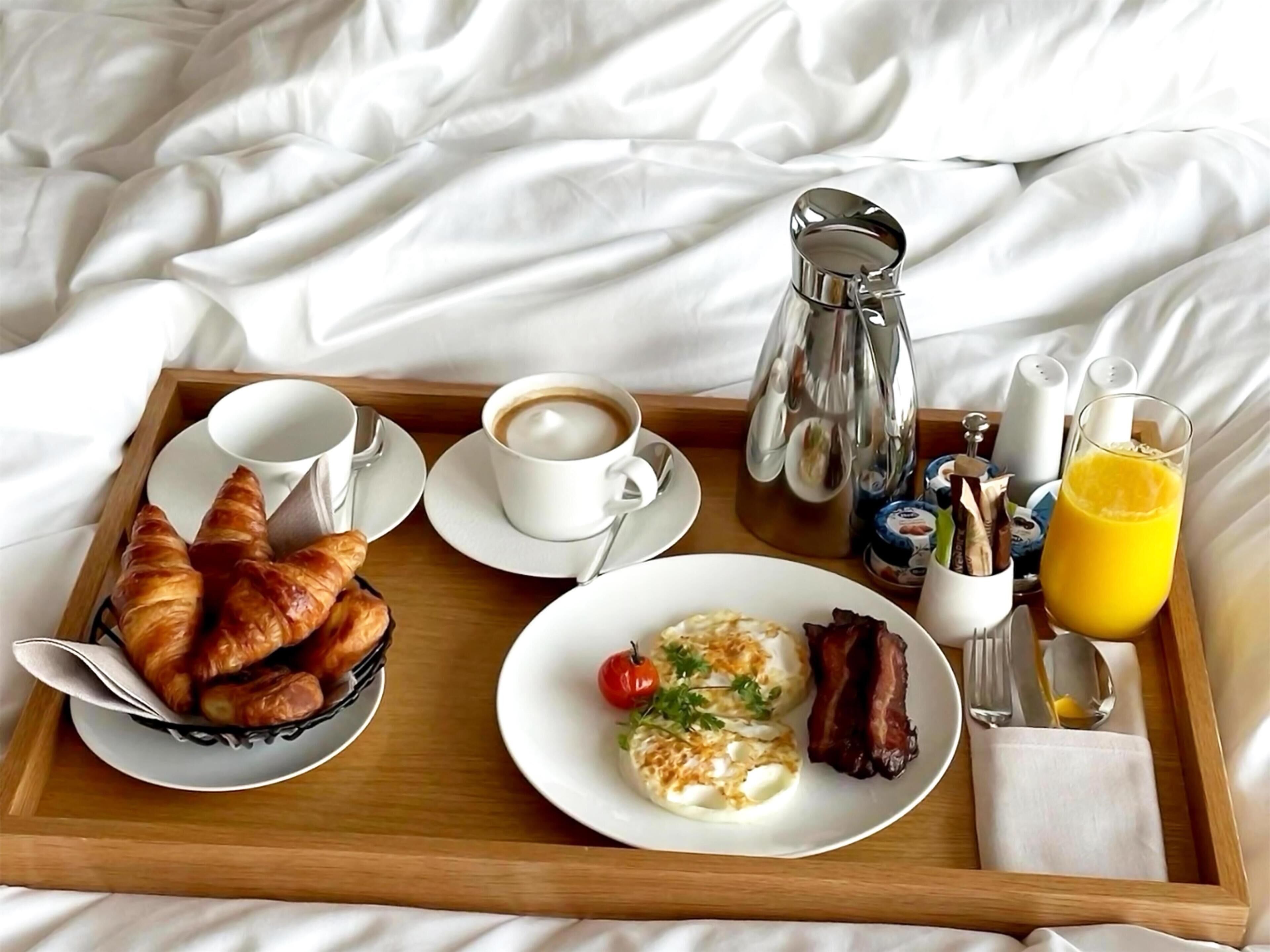 Bandeja de desayuno con cruasanes, café, huevos, tocino y jugo de naranja sobre una cama.
