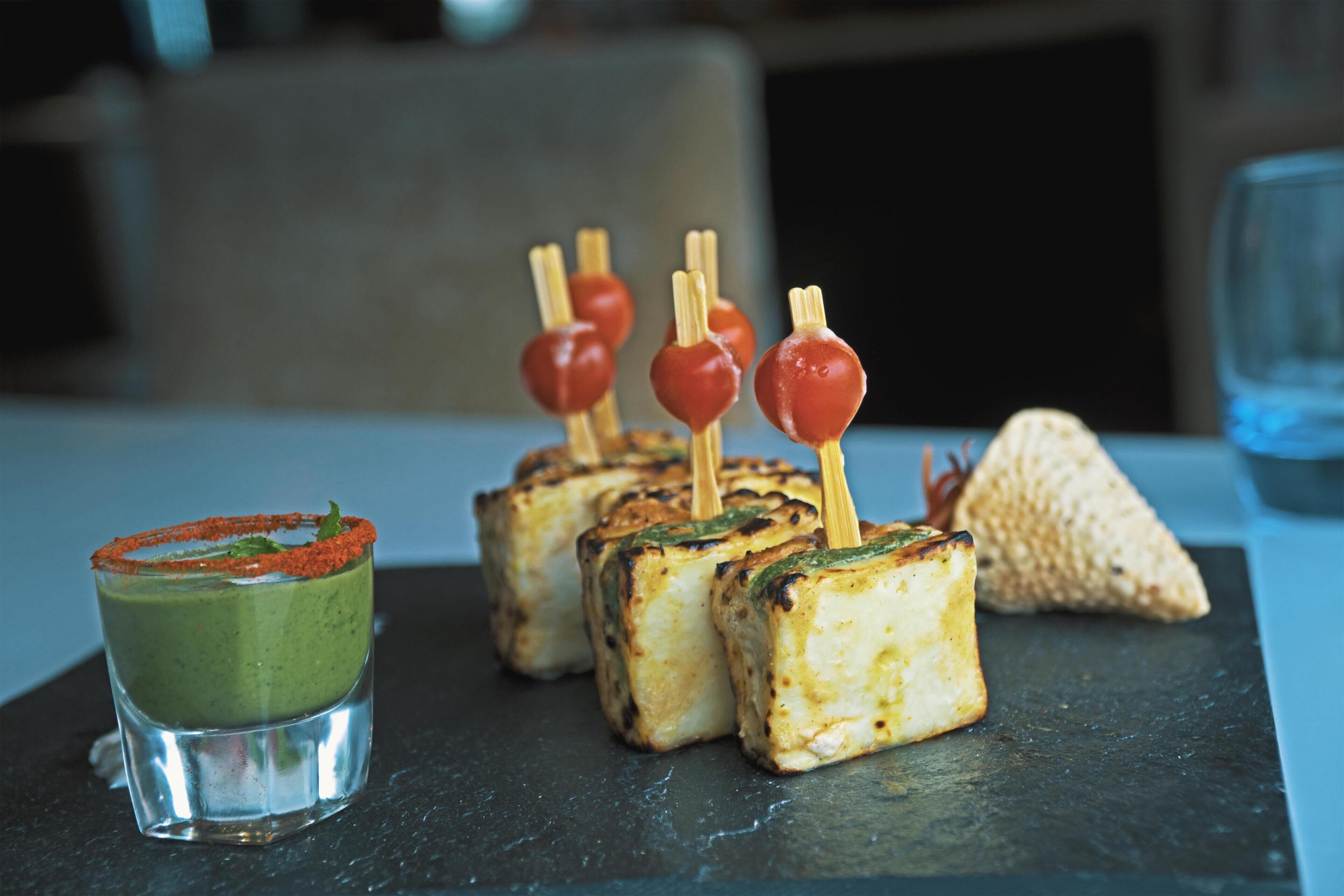 A dessert platter with various items including a green drink, grilled cheese cubes, and a triangular pastry on a slate serving board.