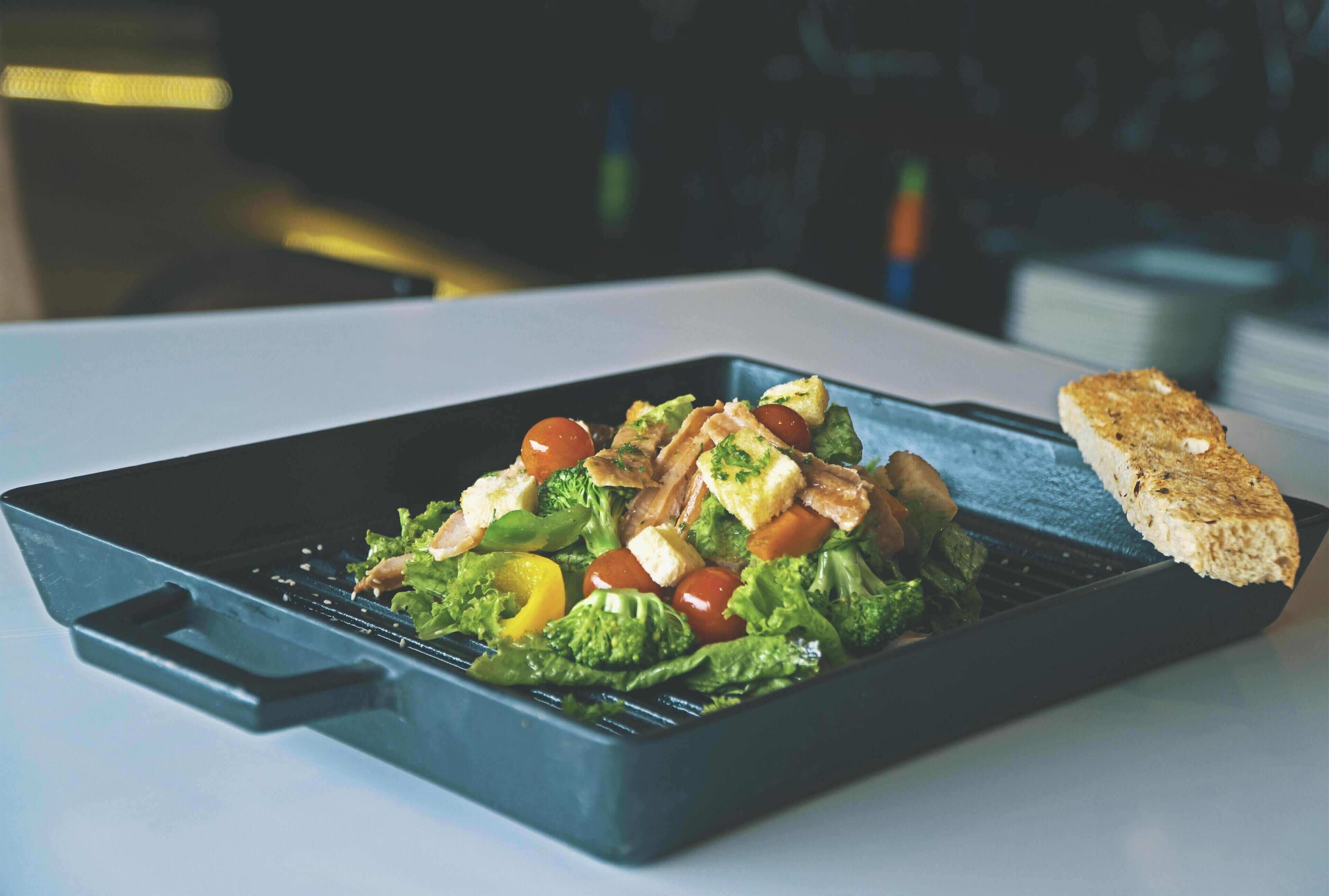 A rectangular metal tray containing a salad with lettuce, tomatoes, and croutons, accompanied by a piece of bread on a white surface.