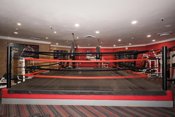 A boxing ring with a red and black color scheme, surrounded by gym equipment and a brick wall.