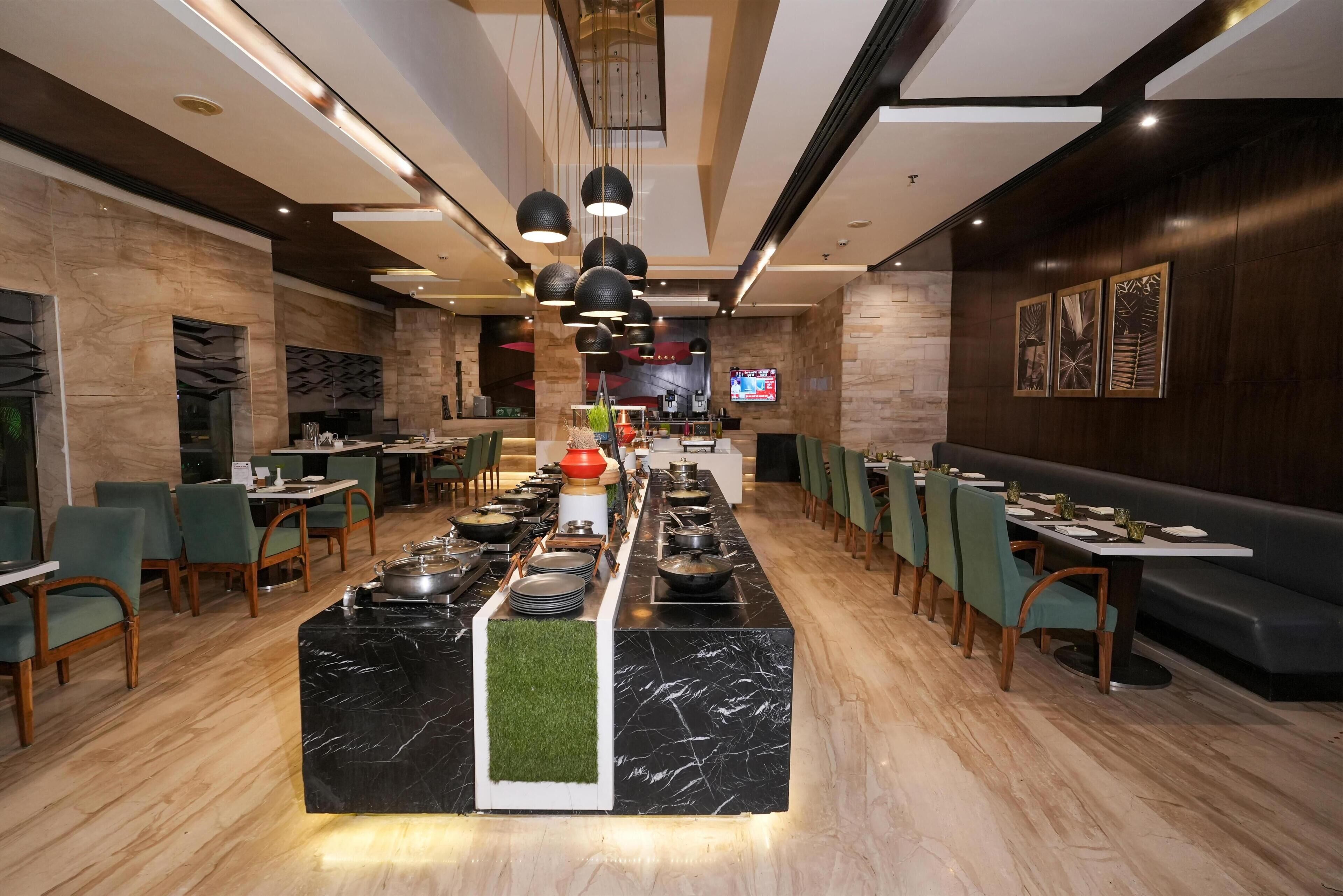 A modern restaurant with a central buffet area, multiple dining tables, and green chairs.