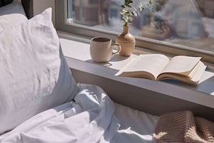 Un lit avec une literie blanche, une fenêtre donnant sur des immeubles, un livre, une tasse et un vase avec une plante.