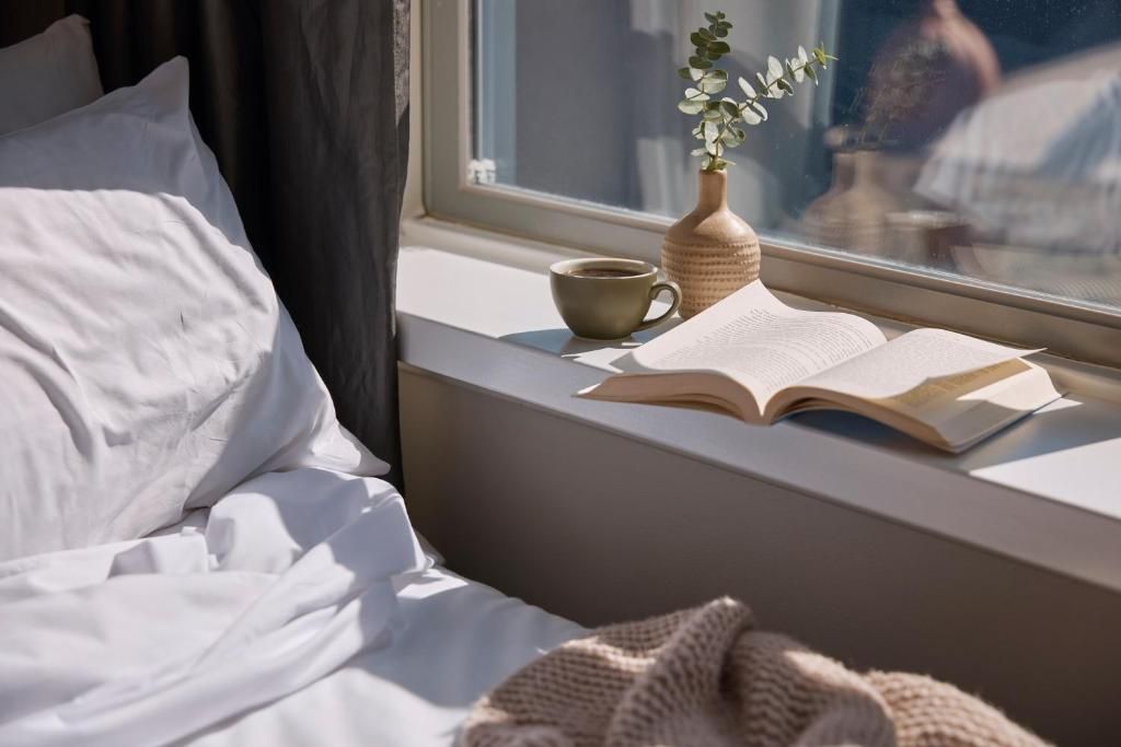 A bed with white linens, a window sill with a book, a cup, and a vase with a plant.