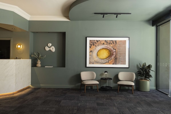 Un espace d’accueil avec un comptoir en marbre, deux chaises, un tableau abstrait encadré, une petite table avec une lampe et une plante en pot.