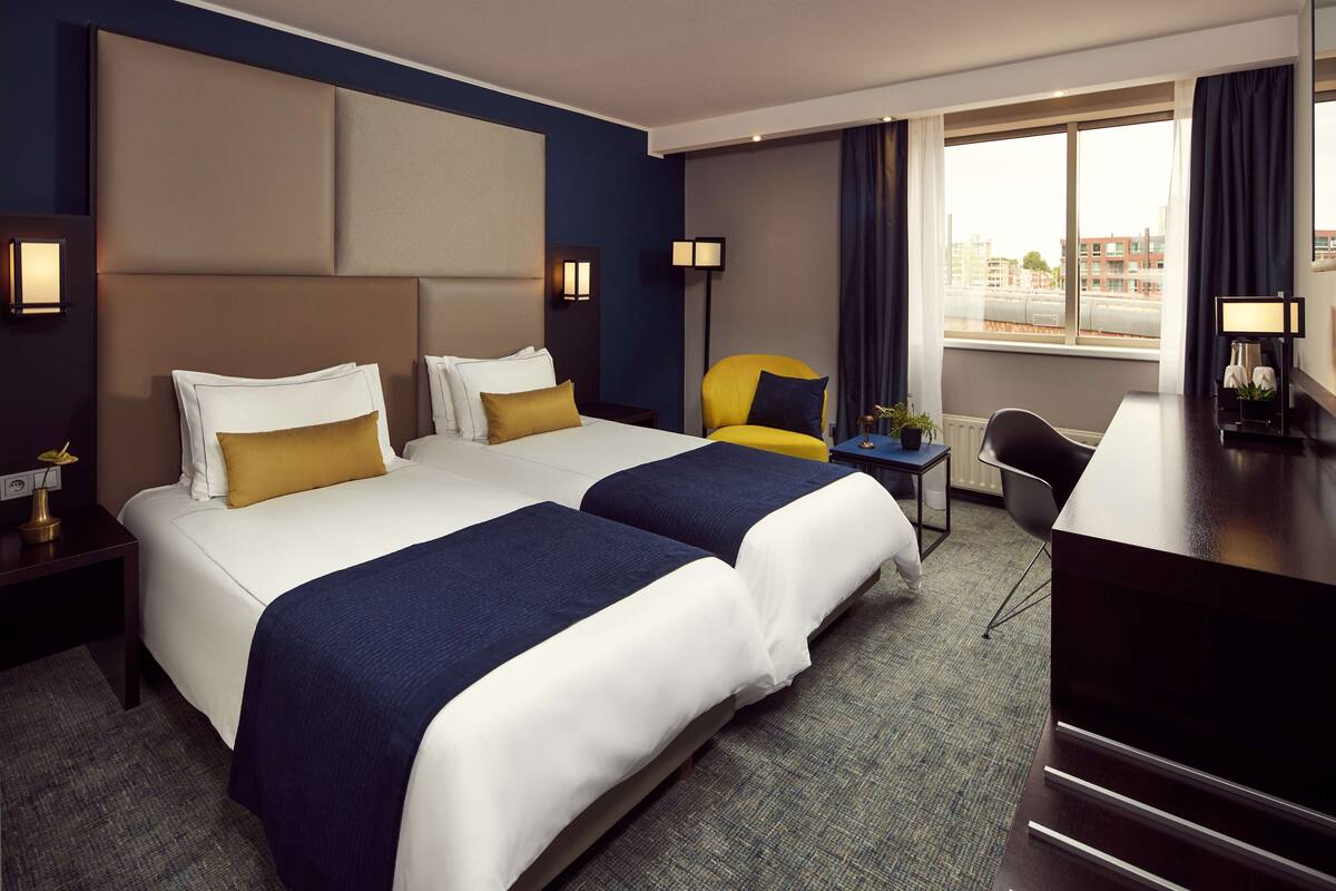 A hotel room with two beds, a desk, a chair, and a view of the city.