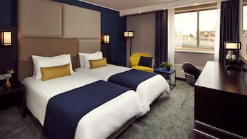 A hotel room with two beds, a desk, a chair, and a view of the city.