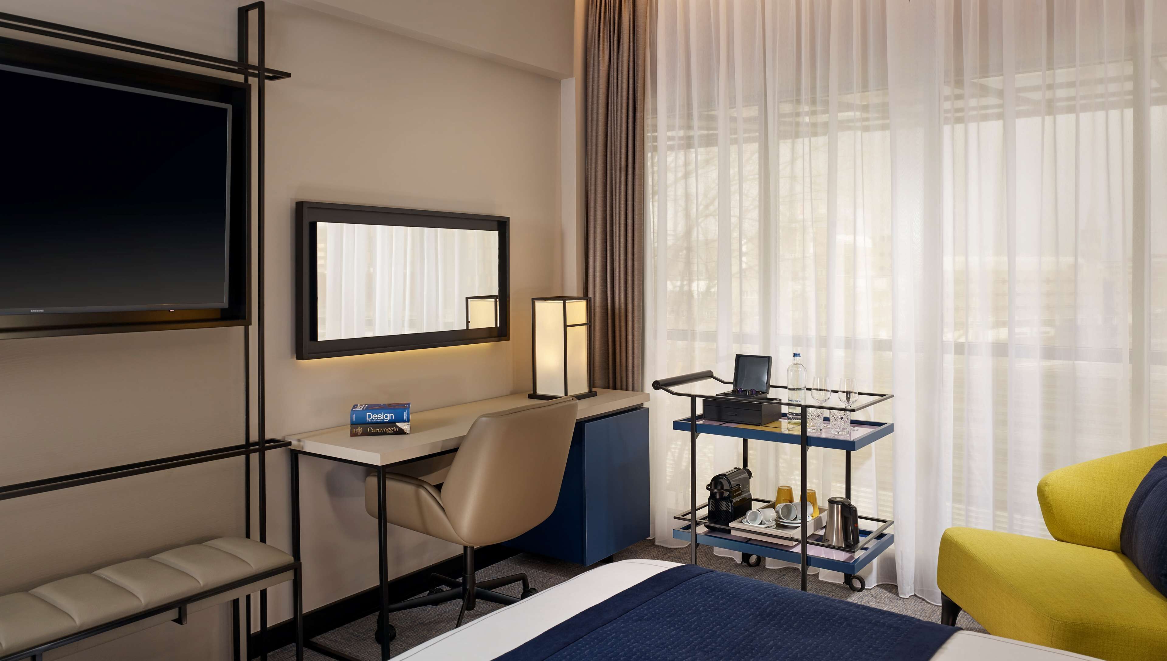 A hotel room with a bed, a desk with a chair, a TV, a bedside table with a lamp, a window with curtains, and a cart with toiletries.