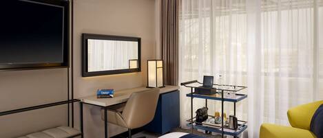 Habitación de hotel con cama, escritorio y silla, televisor, mesita de noche con lámpara, ventana con cortinas y carrito con artículos de aseo.