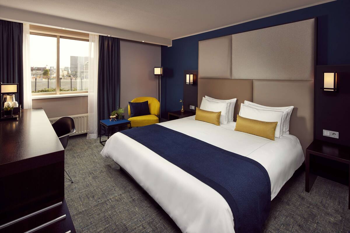A hotel room with a large bed, a desk, a chair, and a window with curtains.
