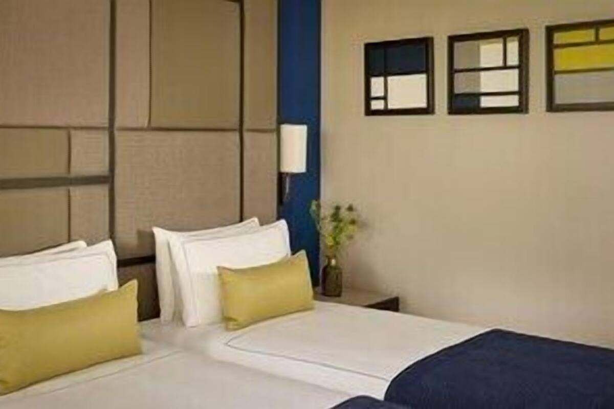 A hotel room with a bed, pillows, a nightstand, a lamp, and a wall with three windows.