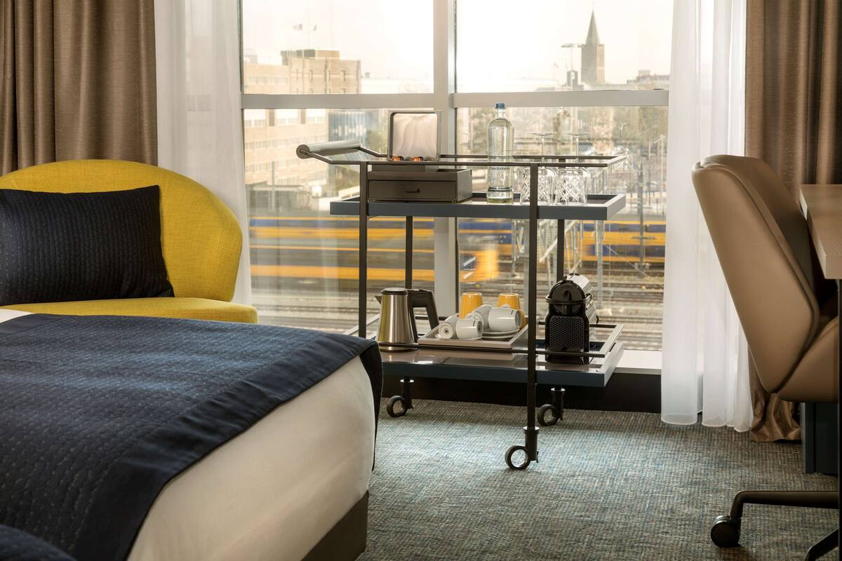 A hotel room with a bed, a yellow armchair, a desk with a cart, and a view of the cityscape.