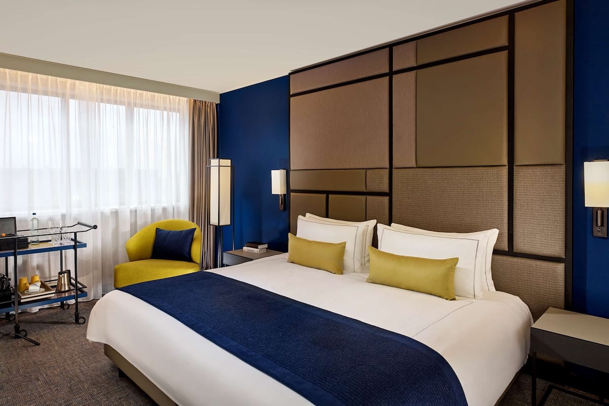 A hotel room with a large bed, a desk with a computer, a chair, and a small table with a lamp.