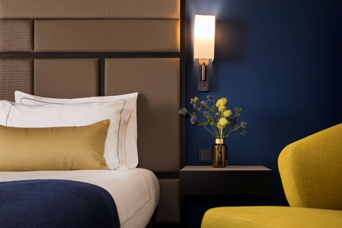 A hotel room with a bed, a nightstand with a lamp, a vase with flowers, and a yellow chair.