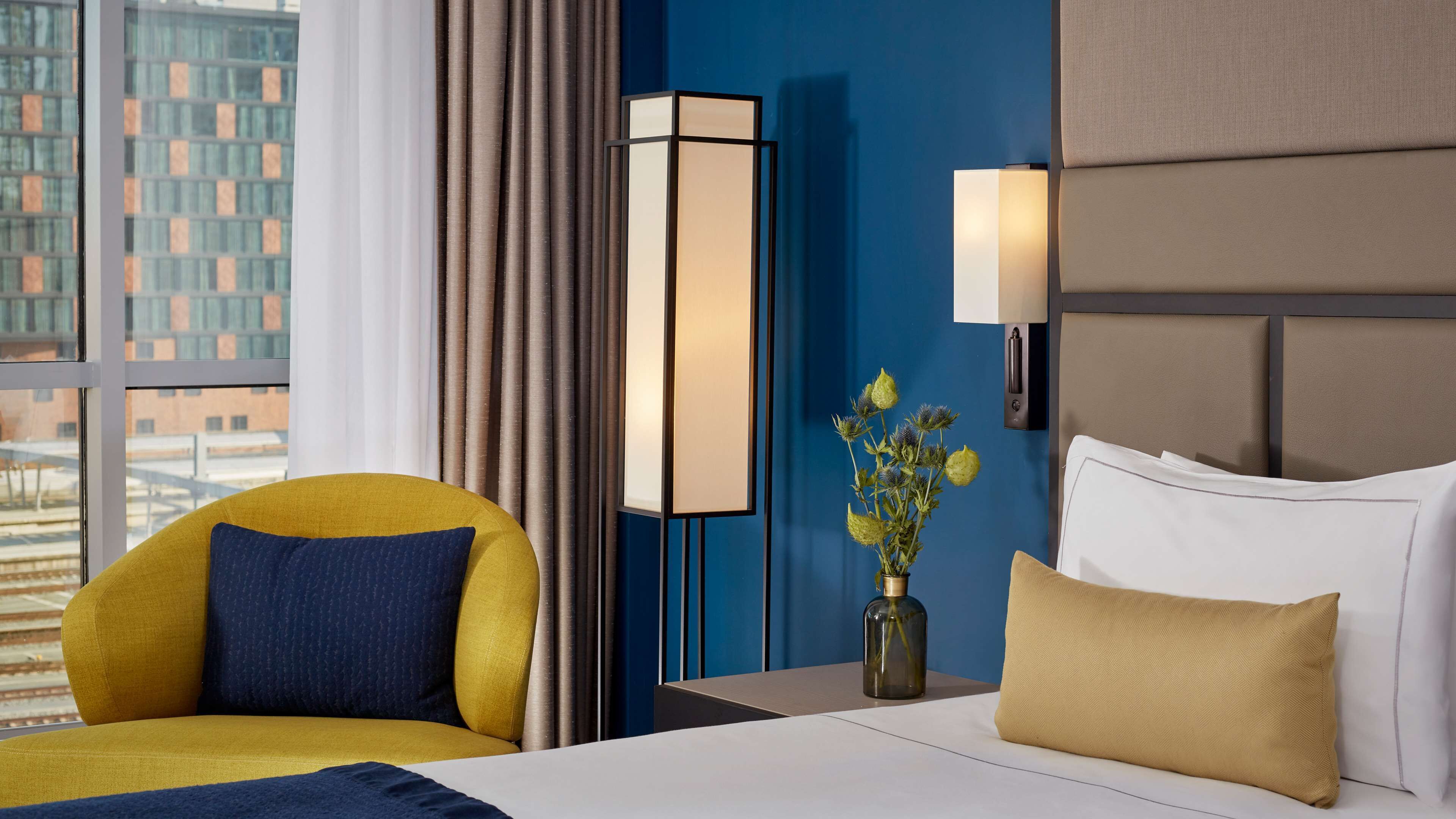 A hotel room with a bed, a yellow armchair, a vase with flowers, a bedside table, and a nightstand.