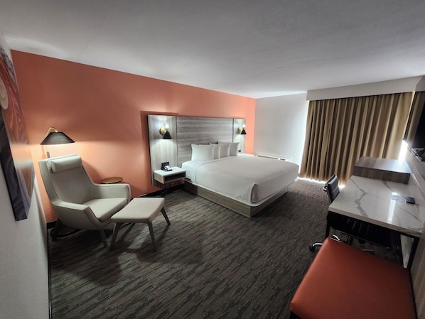 A hotel room with a bed, a desk, a chair, a desk lamp, a bedside table, and a large mirror.