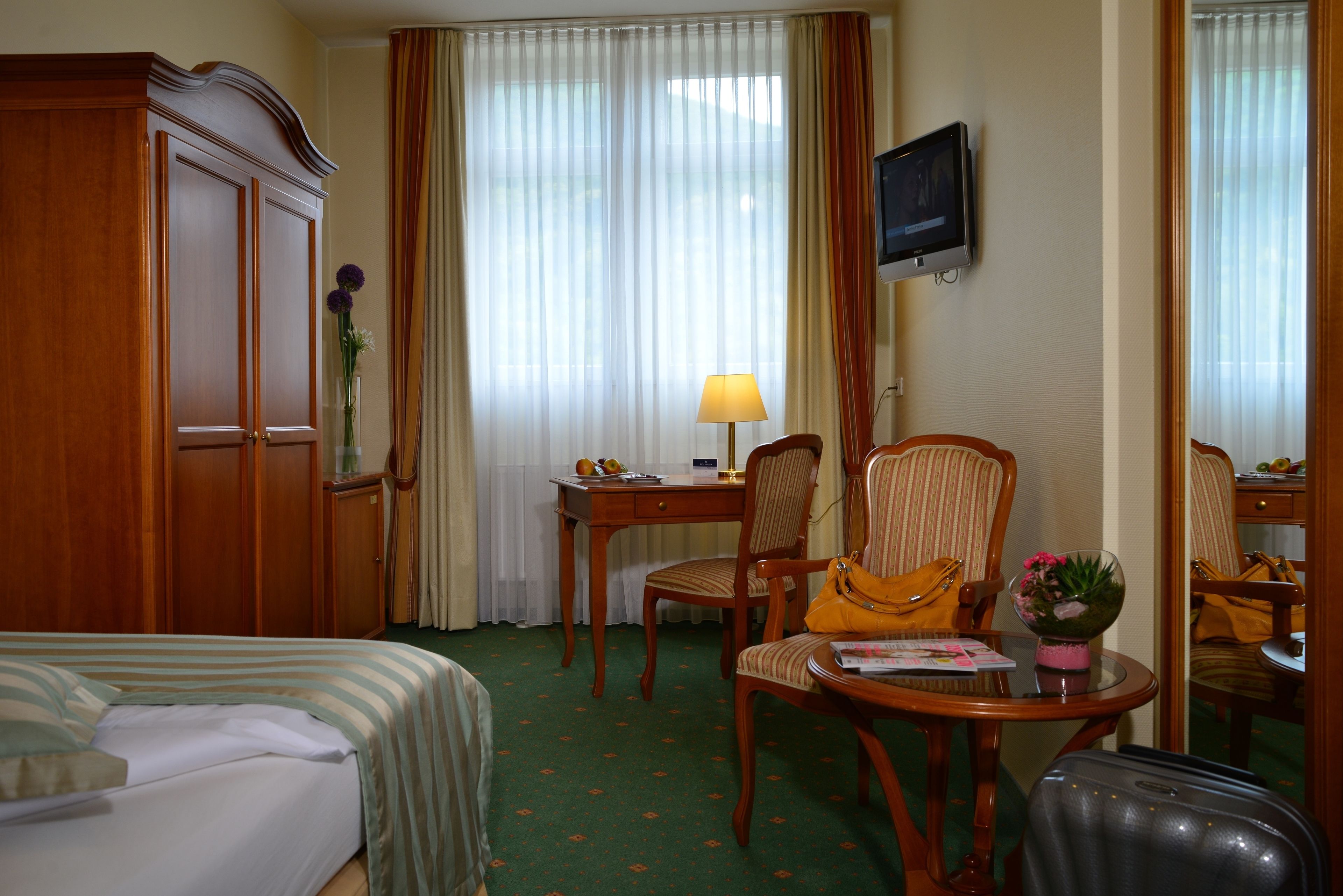 Habitación de hotel con cama, escritorio con lámpara, dos sillas, televisor y maleta.
