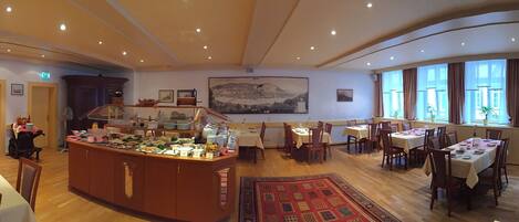 A spacious dining area with multiple tables and chairs, a central buffet counter, and large windows providing natural light.