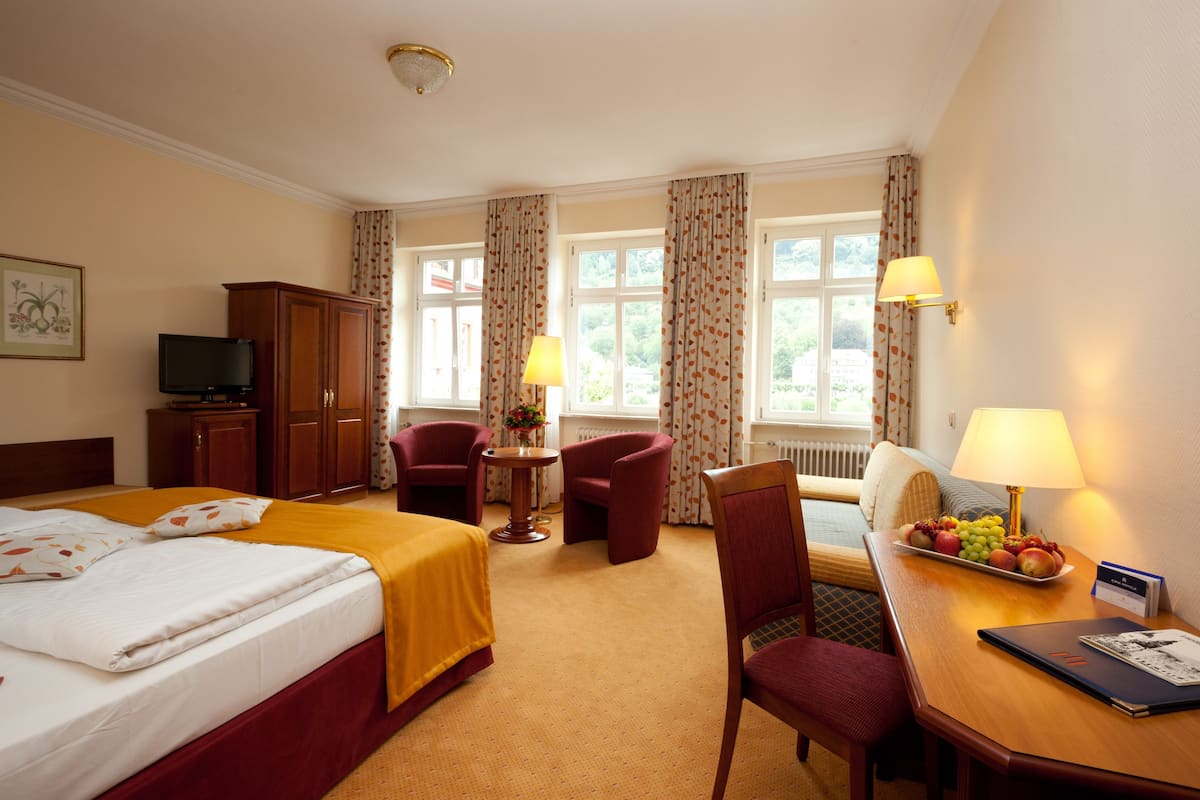 A hotel room with a bed, two armchairs, a desk with a lamp, a television, and a fruit tray.