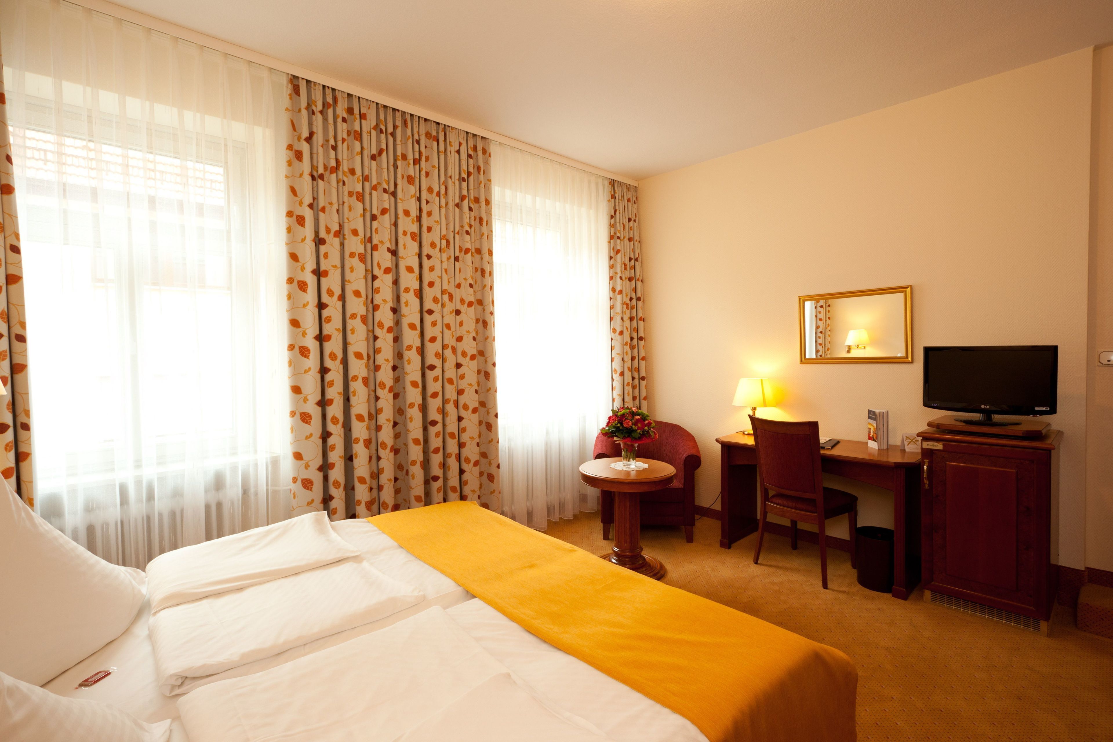 A hotel room with a bed, a desk with a chair, a television, and a small table with a lamp and flowers.